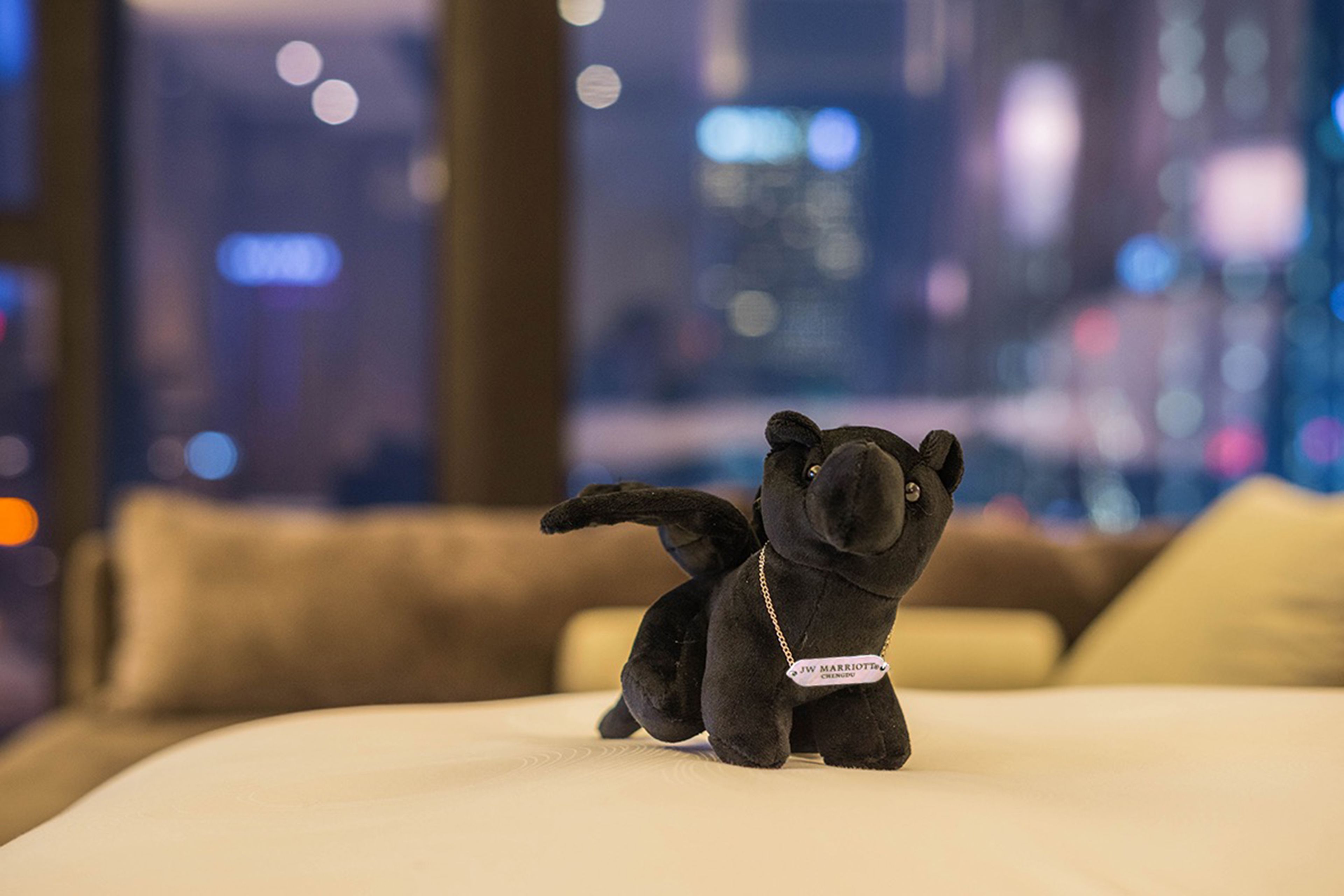 A plush black animal toy on a bed with a blurred cityscape view in the background.