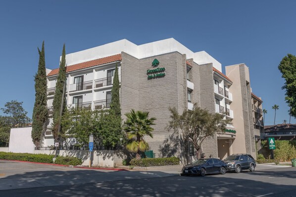 Un hôtel à plusieurs étages, dont la façade est ornée d’un grand logo vert, et qui présente des balcons et un toit recouvert de tuiles rouges.
