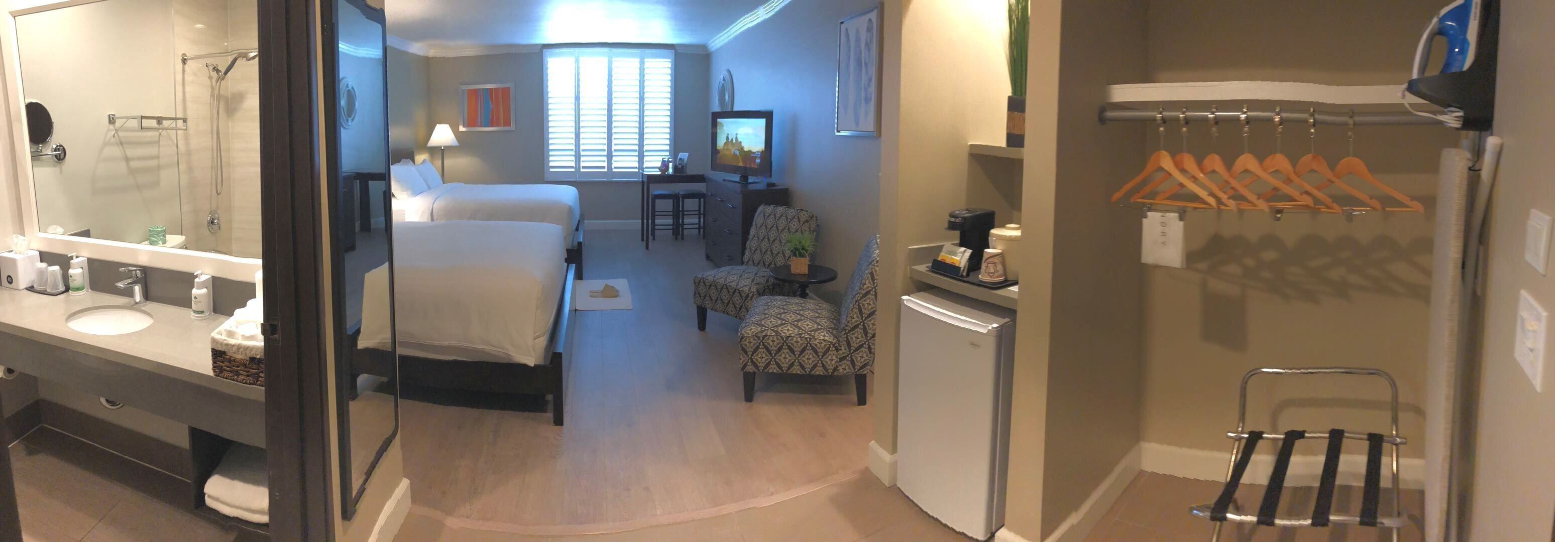 Habitació d'hotel amb llit, escriptori, cadira, nevera petita, mirall, bany amb rentapel i dutxa, i finestra amb persianes.