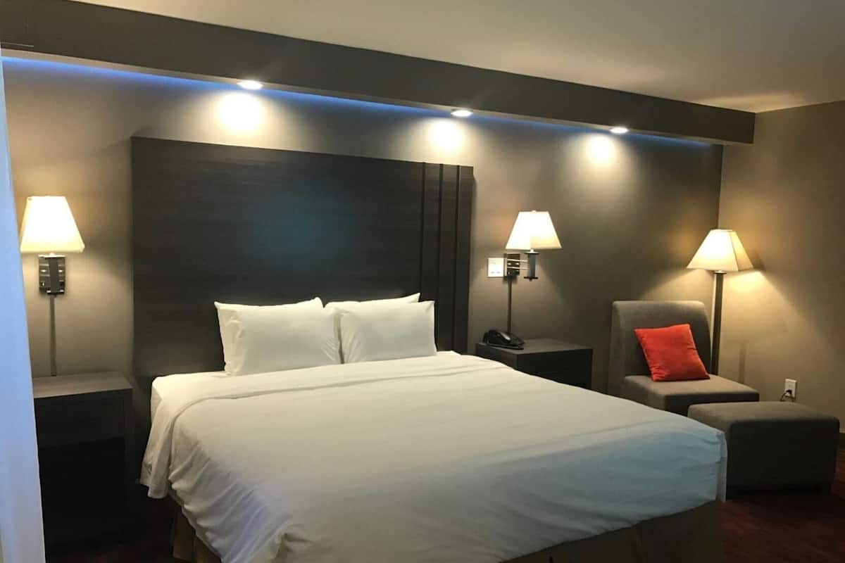 A hotel room with a large bed, two bedside tables with lamps, a chair with a red pillow, and a small ottoman.
