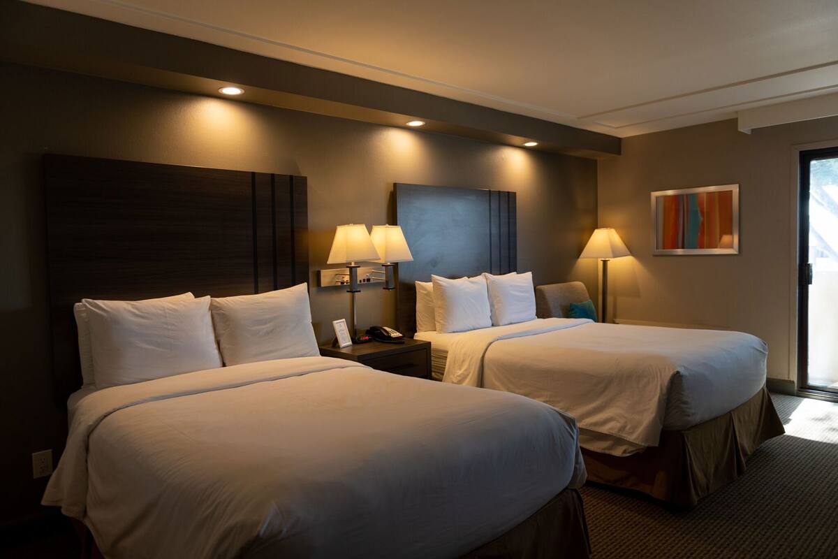 A hotel room with two beds, bedside lamps, a phone, a painting, and a view of the outdoors.