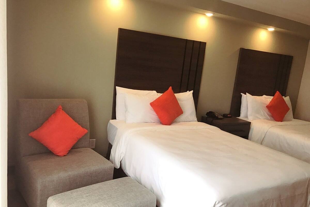 A hotel room with two beds, each with white bedding and red pillows, a grey armchair, and a small table with a phone.