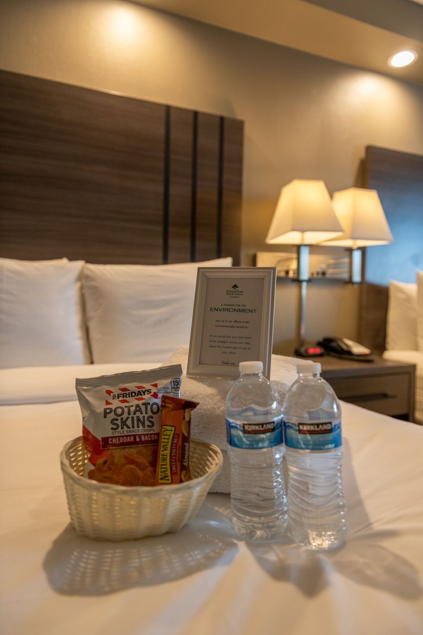 Habitación de hotel con una cama, un cartel sobre el medio ambiente, un bol de aperitivos y dos botellas de agua.