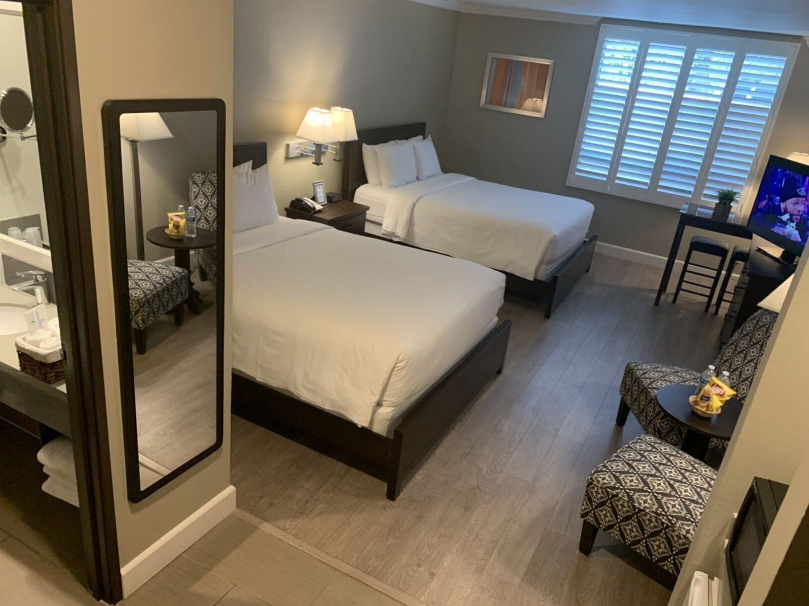 A hotel room with two beds, a desk, a chair, a TV, and a window with blinds.