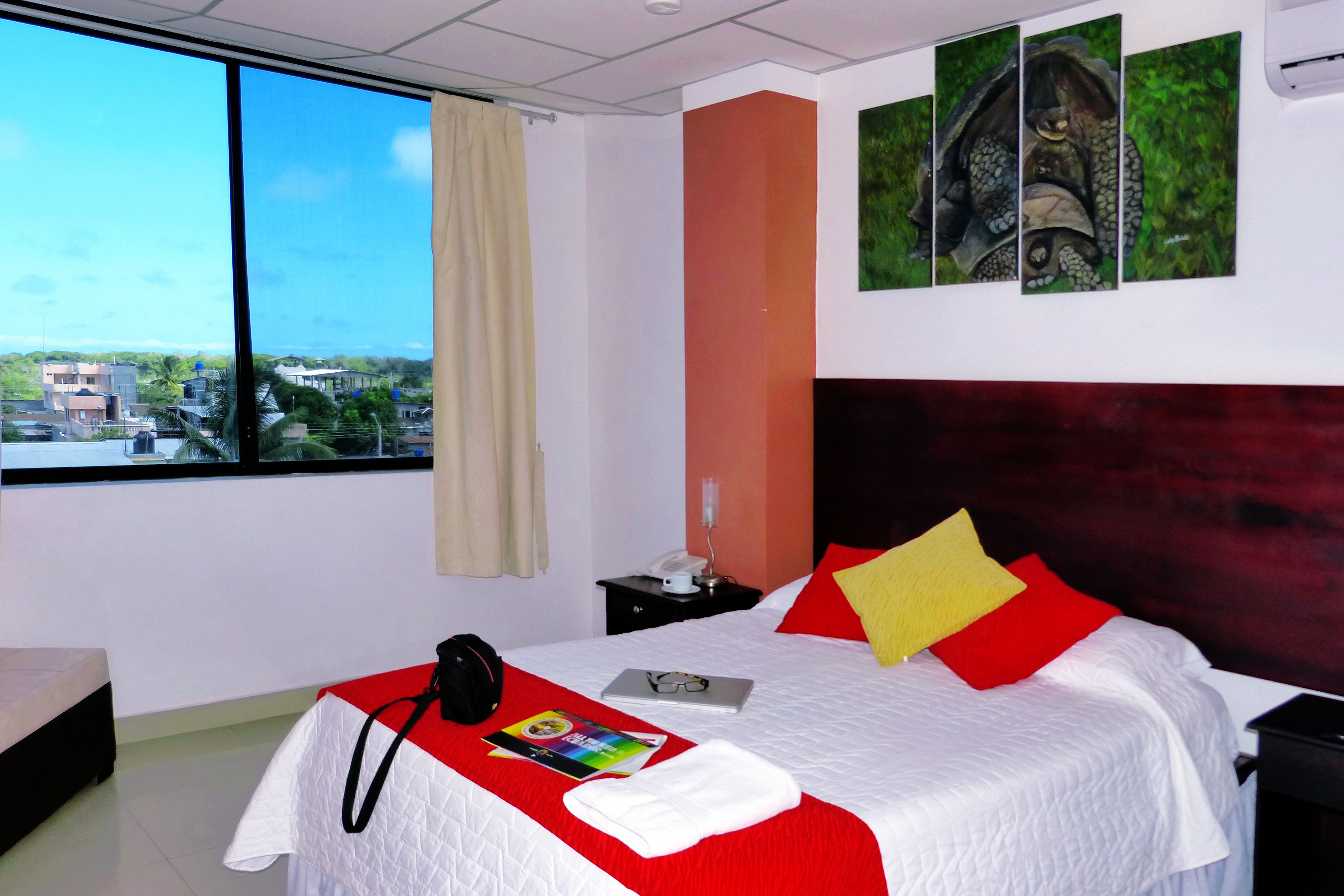 A hotel room with a large window, a bed with red and yellow pillows, a laptop, a pair of glasses, and a book on the bed.