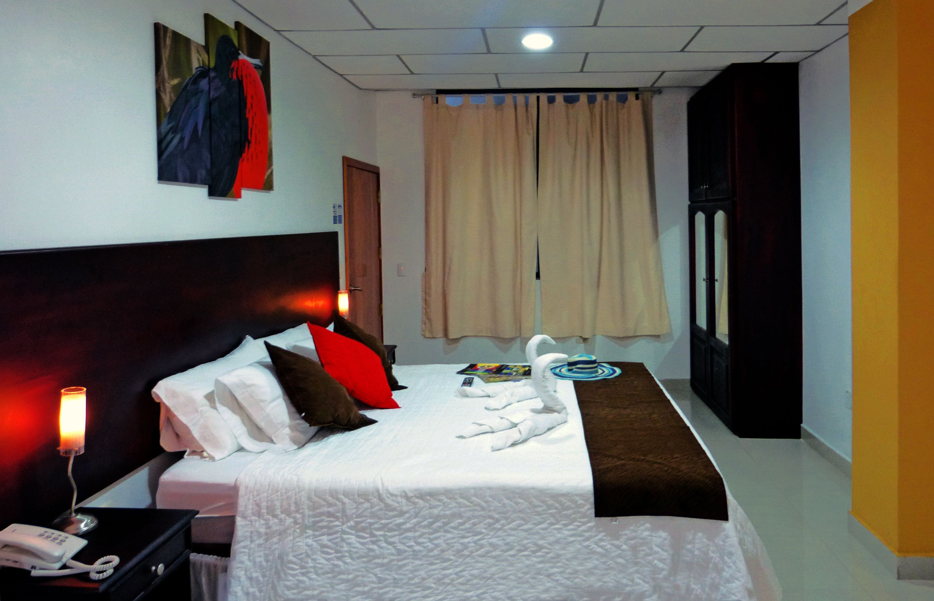 Habitación de hotel con cama, mesita de noche con lámpara, teléfono, espejo, ventana con cortinas y una obra de arte colgada en la pared.
