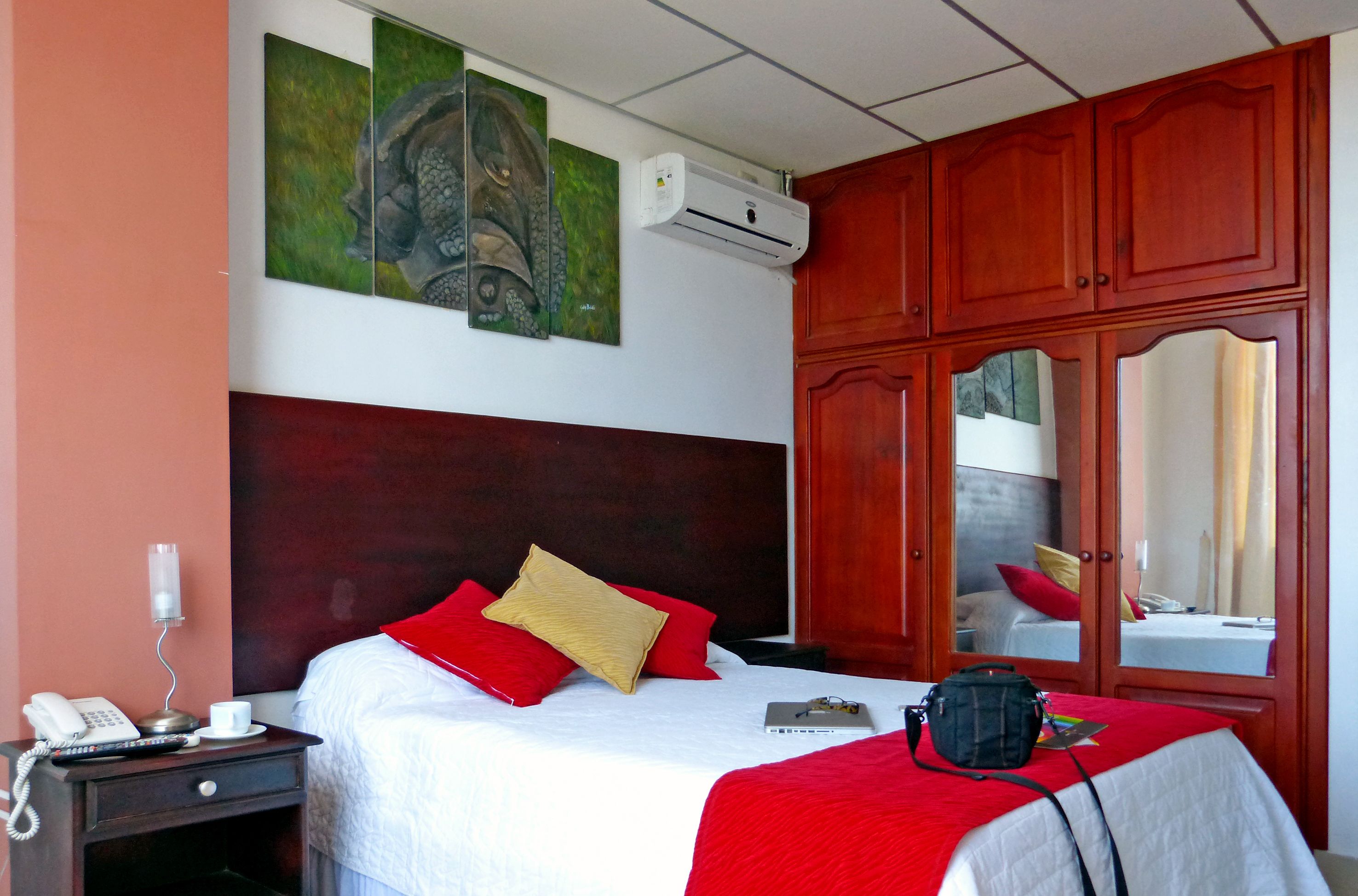 A hotel room with a bed, a bedside table with a phone and a cup, a red and white bedspread, a red and white throw blanket, a black bag on the bed, and a wall-mounted air conditioner.