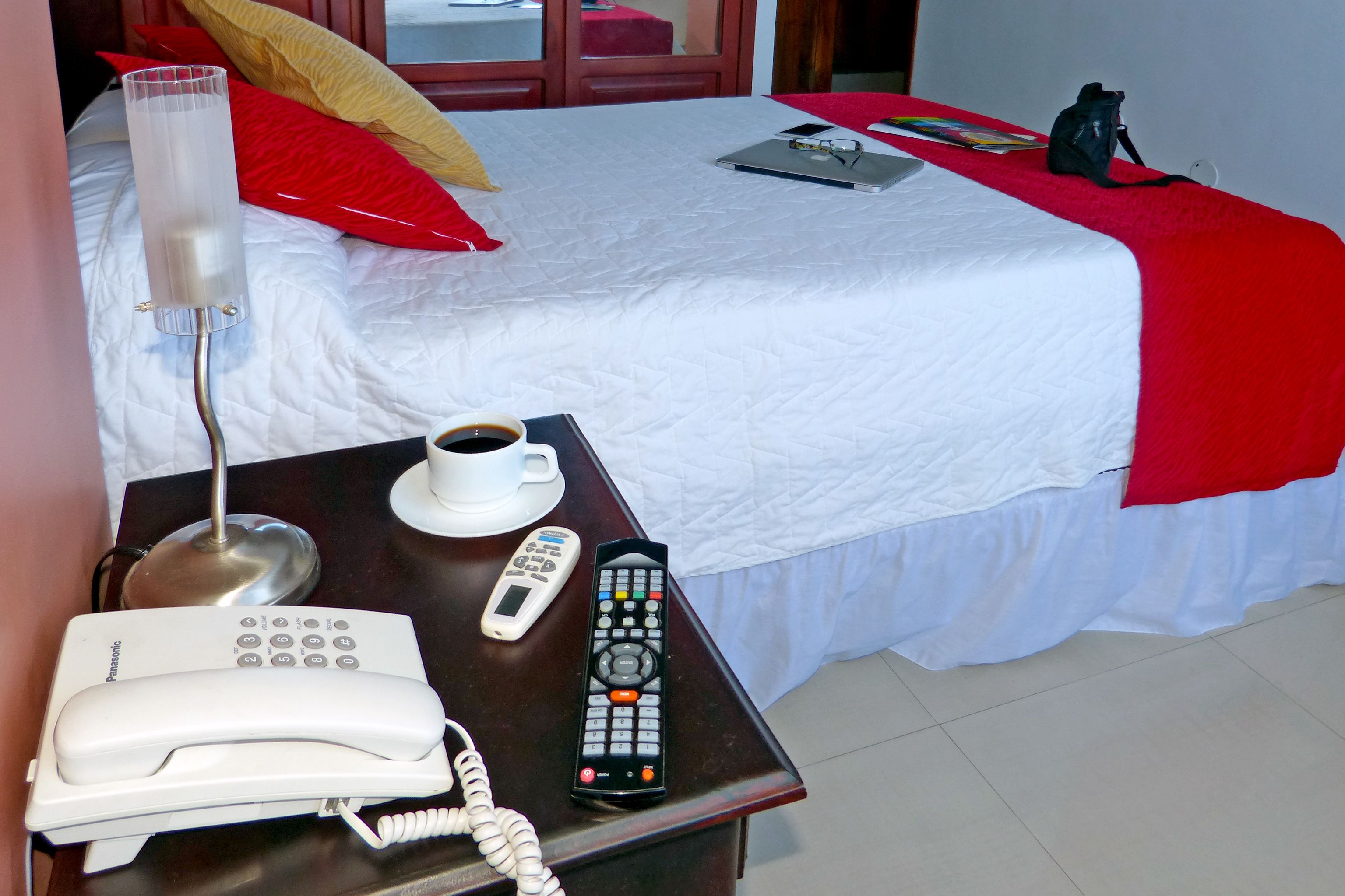 A hotel room with a bed, a bedside table, a lamp, a phone, a remote control, and a cup of coffee.