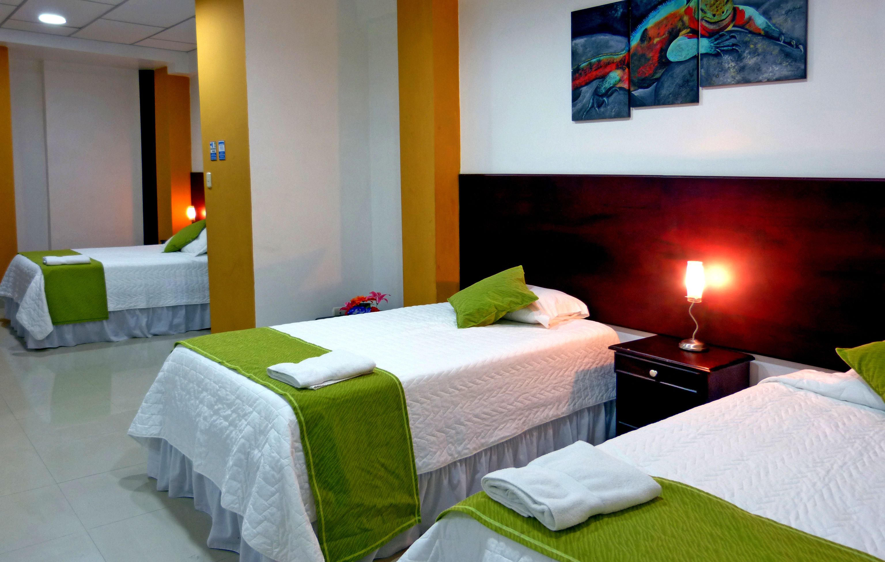 Quarto de hotel com duas camas, cada uma com roupa de cama branca e detalhes verdes, uma mesa de cabeceira com abajur e um quadro na parede.