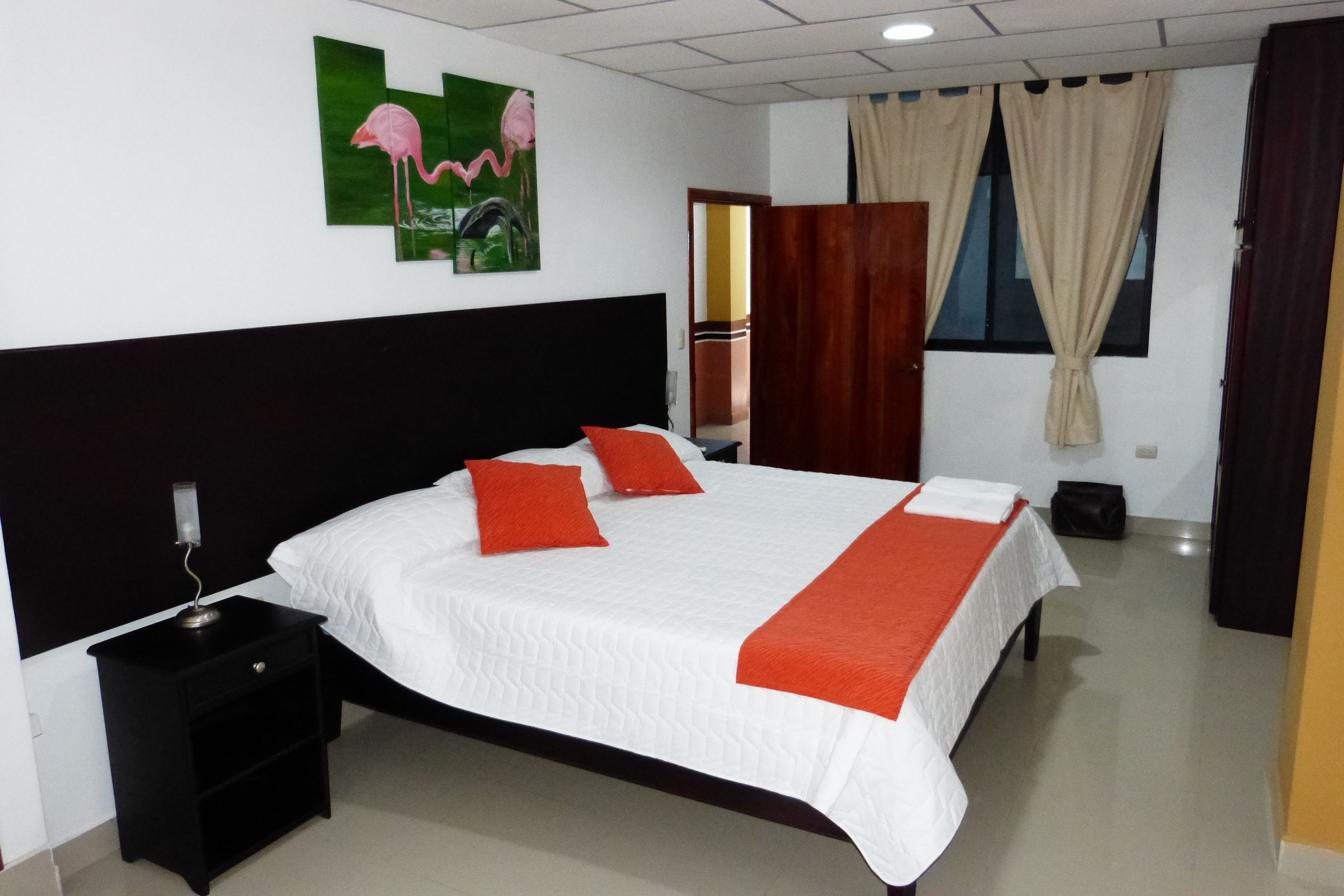 A neatly made bed with white and orange bedding, a black nightstand with a lamp, and a painting of flamingos on the wall.