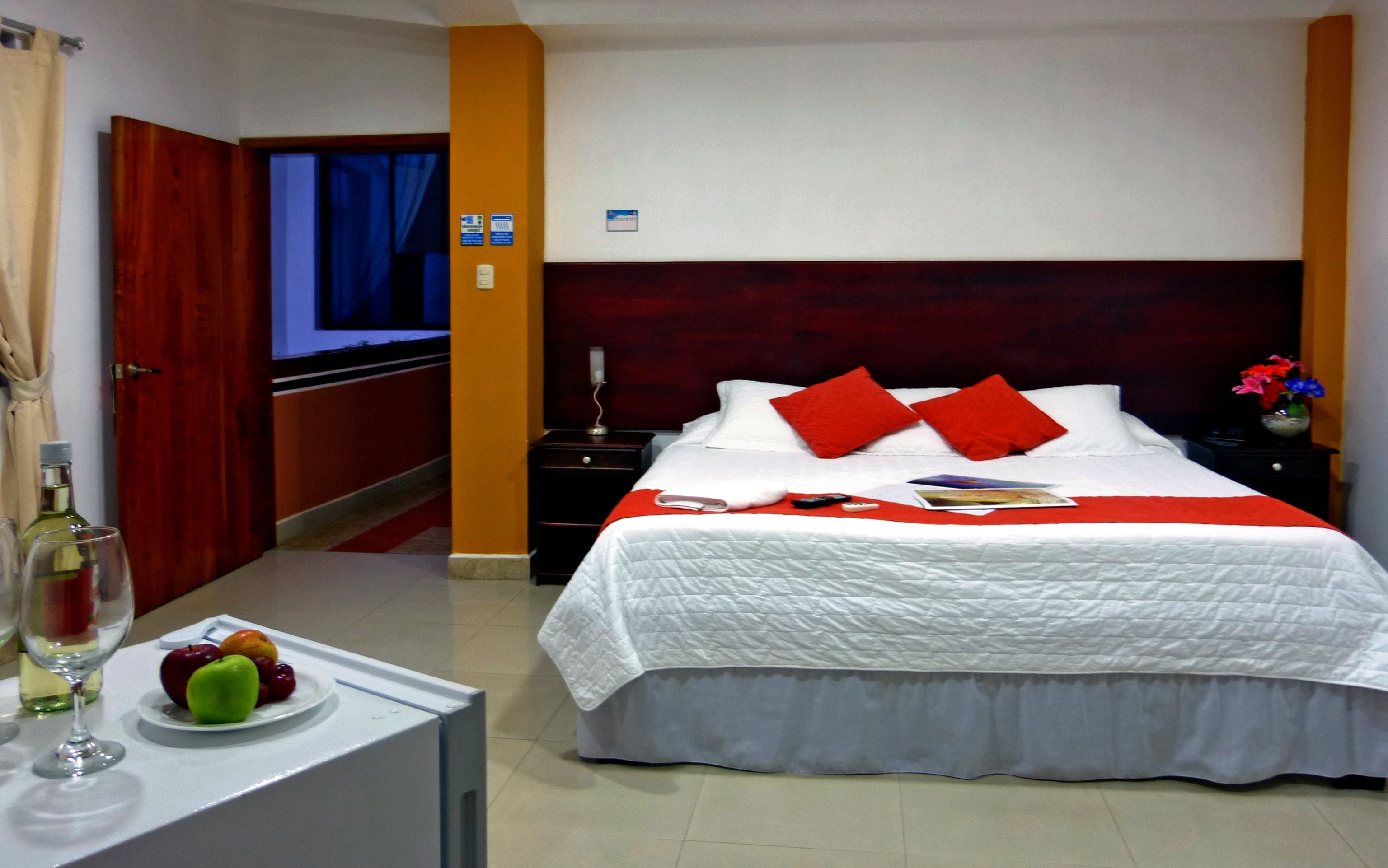 Habitación de hotel con una cama grande, una mesita de noche con una copa de vino y fruta, y vistas al pasillo.