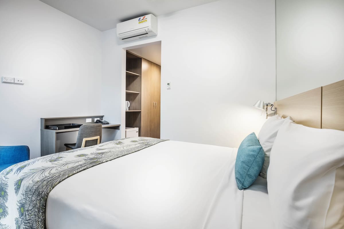 A hotel room with a bed, a desk, a chair, a wardrobe, and an air conditioning unit.