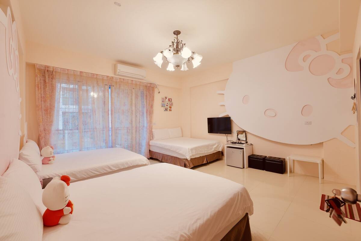 Habitación de hotel con dos camas, un escritorio con una computadora, un televisor y una decoración de Hello Kitty en la pared.