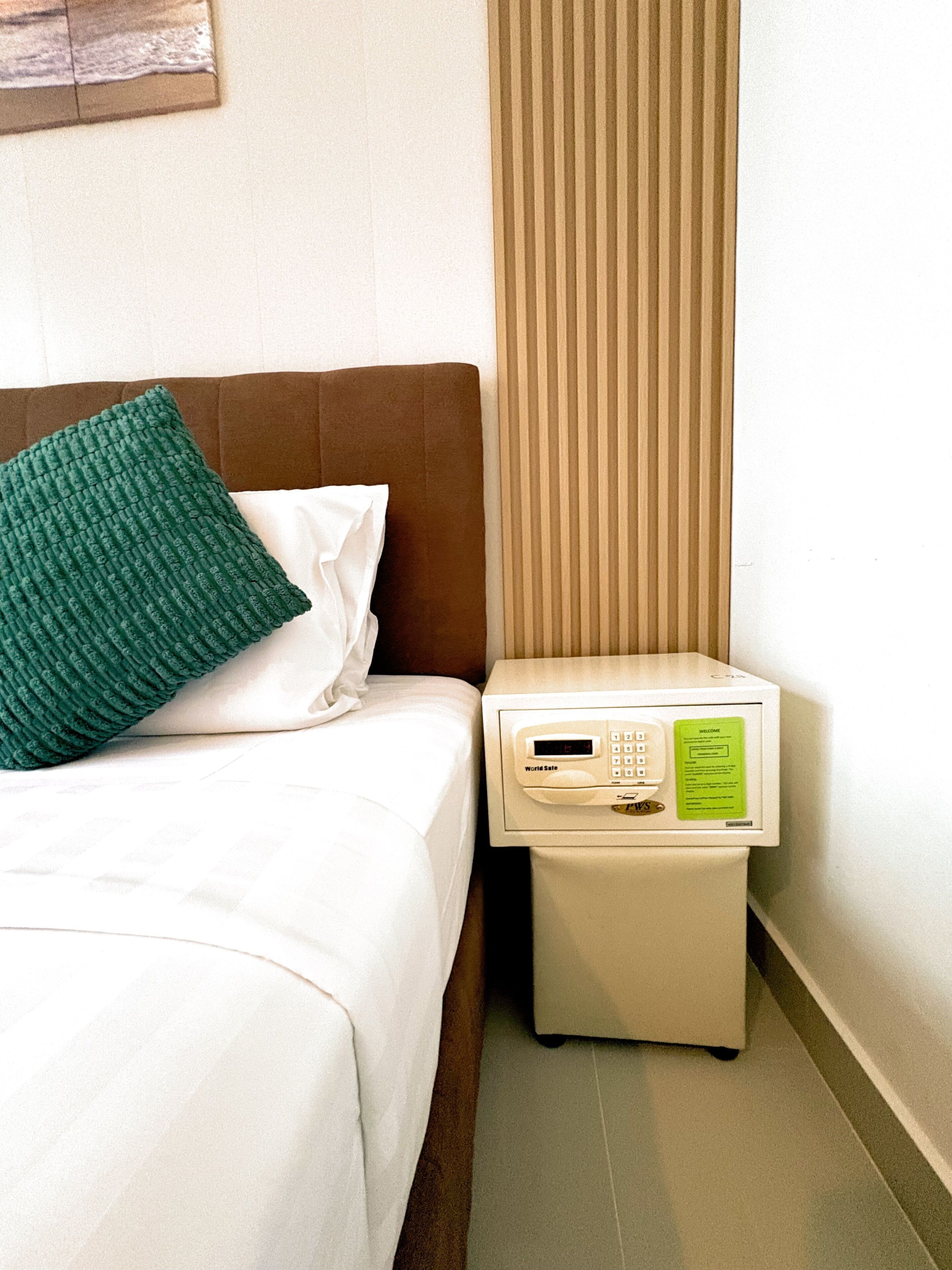 A hotel room with a bed, a green pillow, a nightstand with a digital clock, and a window with blinds.