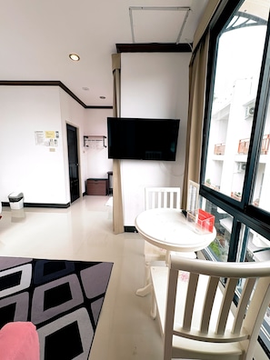 A hotel room with a flat-screen TV mounted on the wall, a round dining table with a chair, a small white table with a red box, and a view of a building through a large window. - Kata Beachwalk Hotel and Bungalows (Karon)