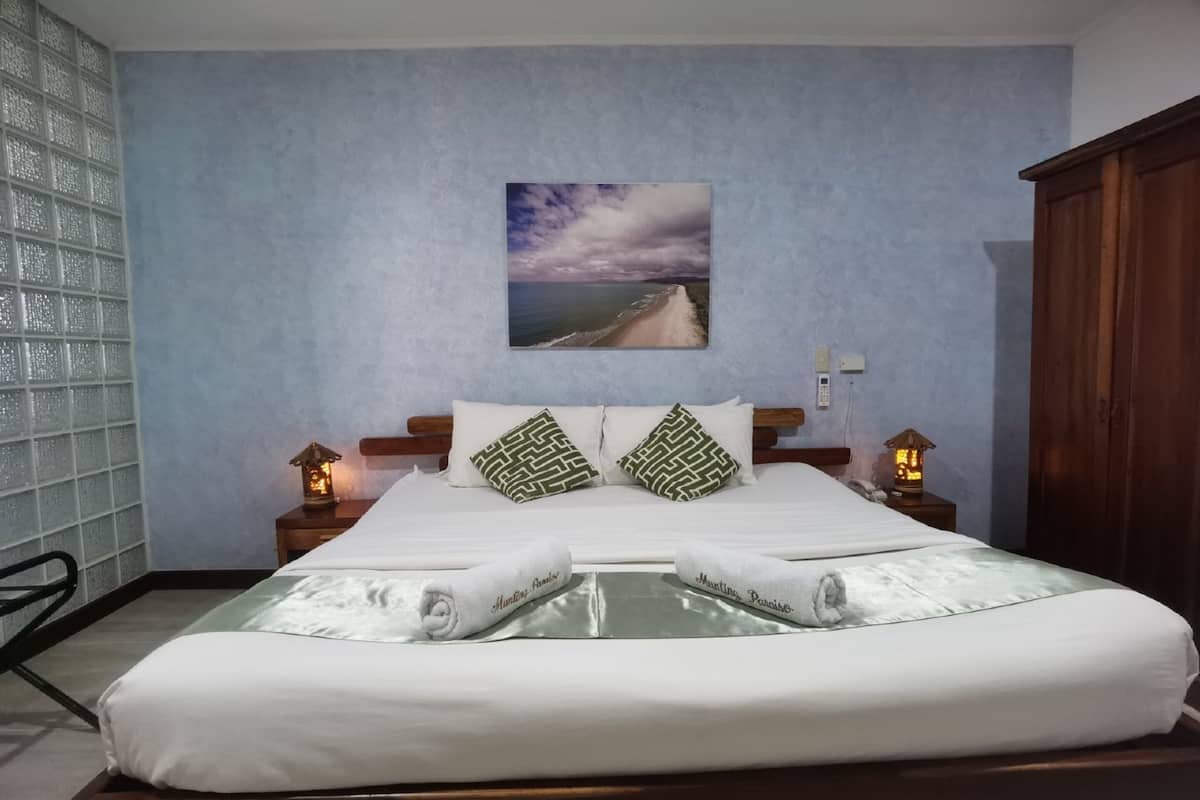 Una cama doble con sábanas blancas, dos almohadones decorativos, un cuadro con una escena de playa y dos lámparas de noche.