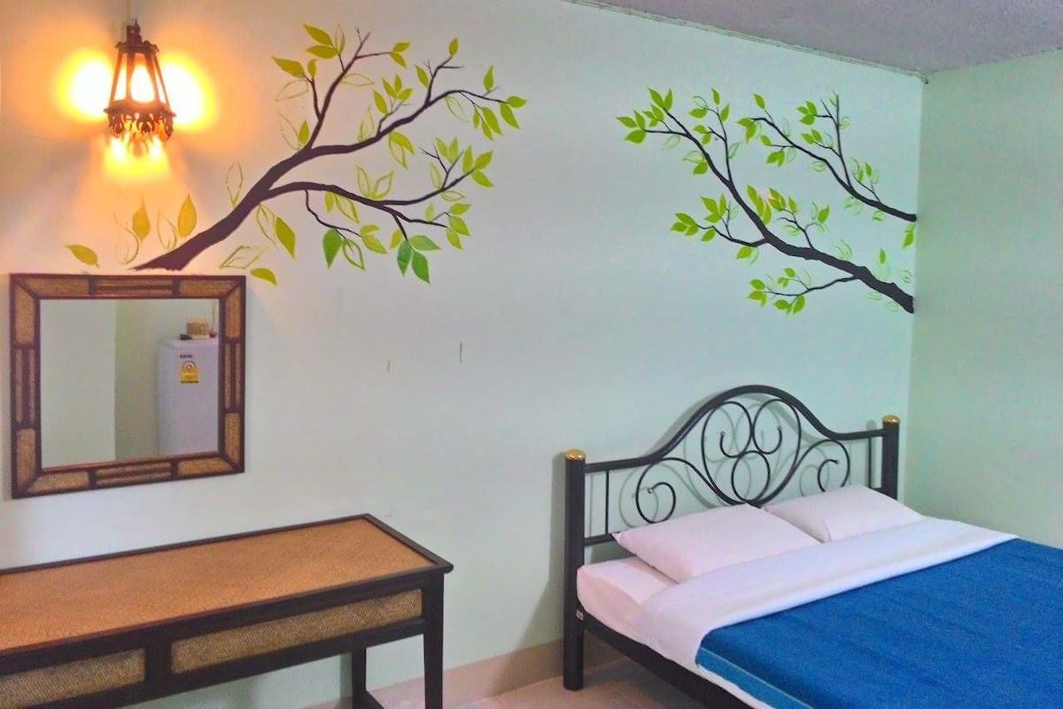 Un dormitorio con cama, mesita de noche, espejo y un cuadro con árboles.