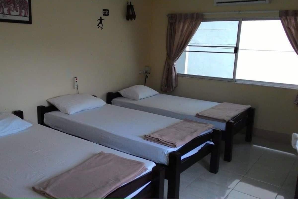 Habitación con cuatro camas individuales alineadas, cada una con almohada y manta doblada. Se ve una ventana con cortinas, un aire acondicionado de pared y una pequeña decoración en la pared.