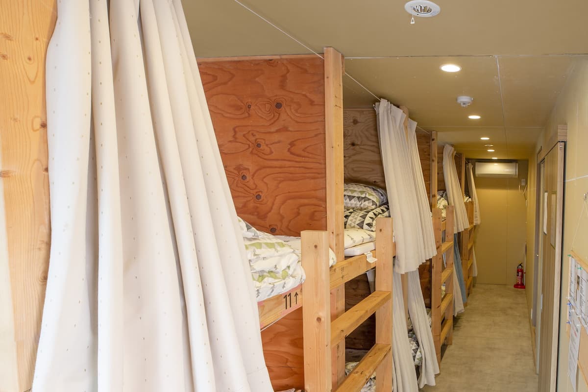 A row of bunk beds with wooden frames and white bedding, a hallway with a carpeted floor, and a door at the end.