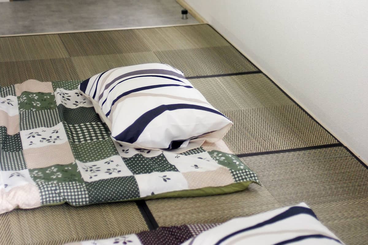 A room with a wooden door, a window with blinds, and a bed with a quilt and pillows.