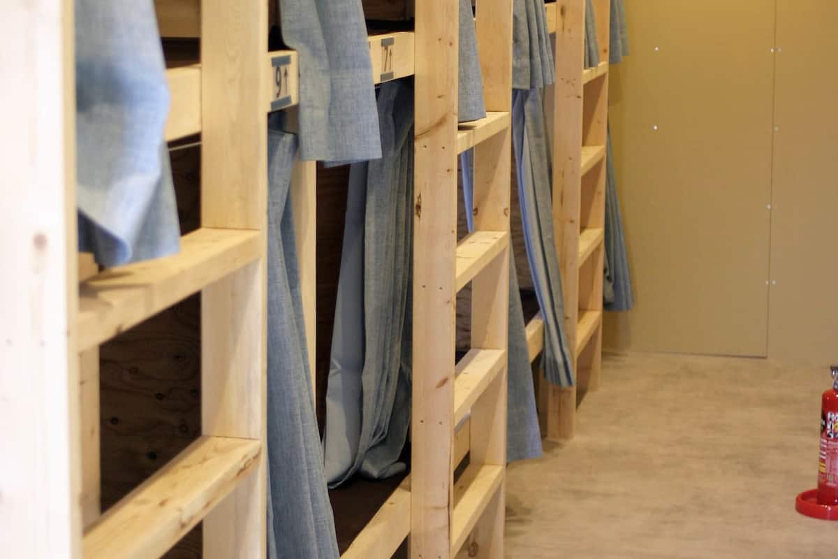 Bunk beds with wooden frames and blue linens, a fire extinguisher, and a beige wall with a recessed light.