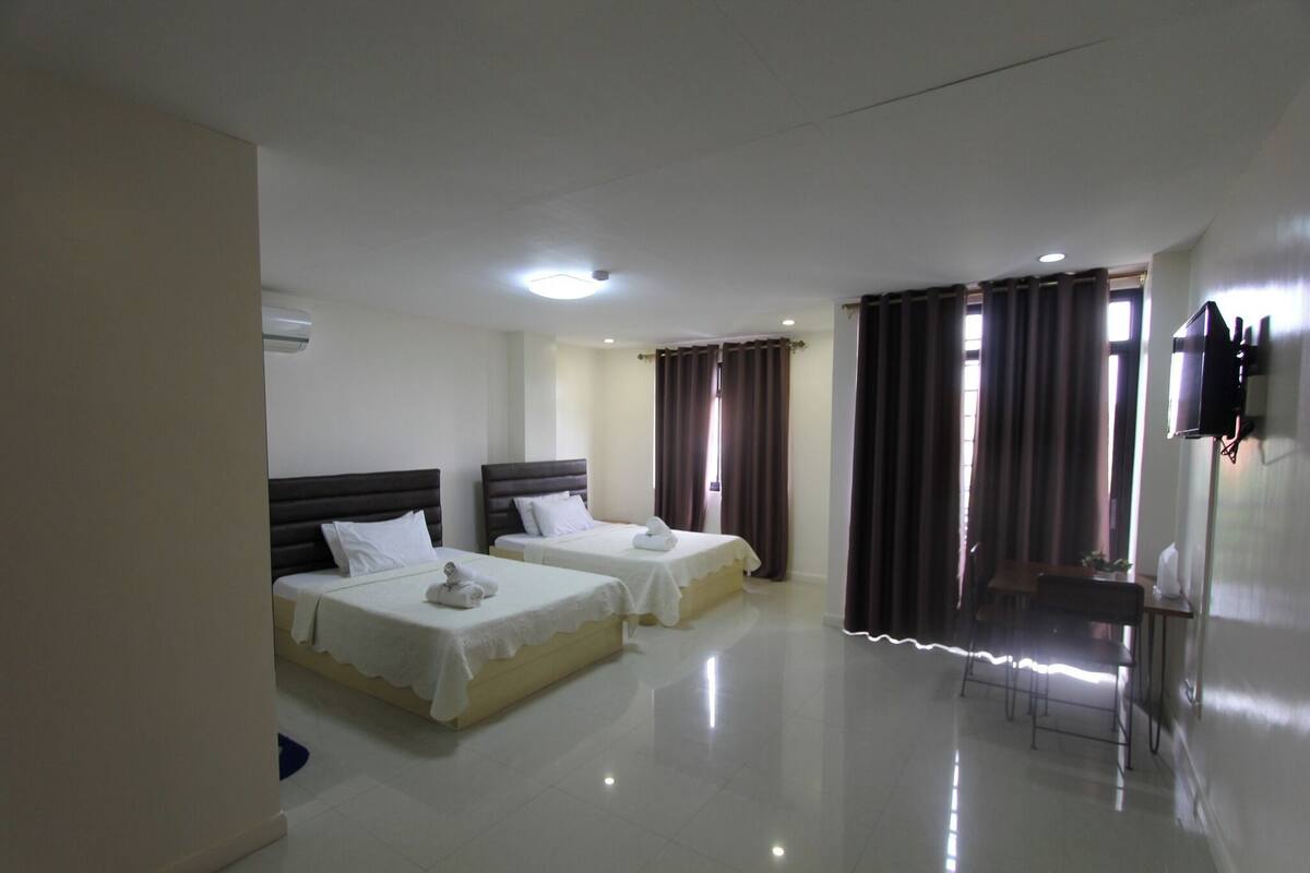 A hotel room with two beds, a desk, a chair, a TV, and an air conditioning unit.