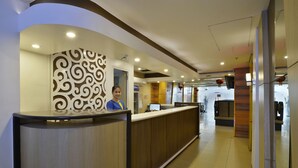 Zona de recepción de hotel con un mostrador de madera, un muro decorativo y un área de descanso con sofá y sillones.