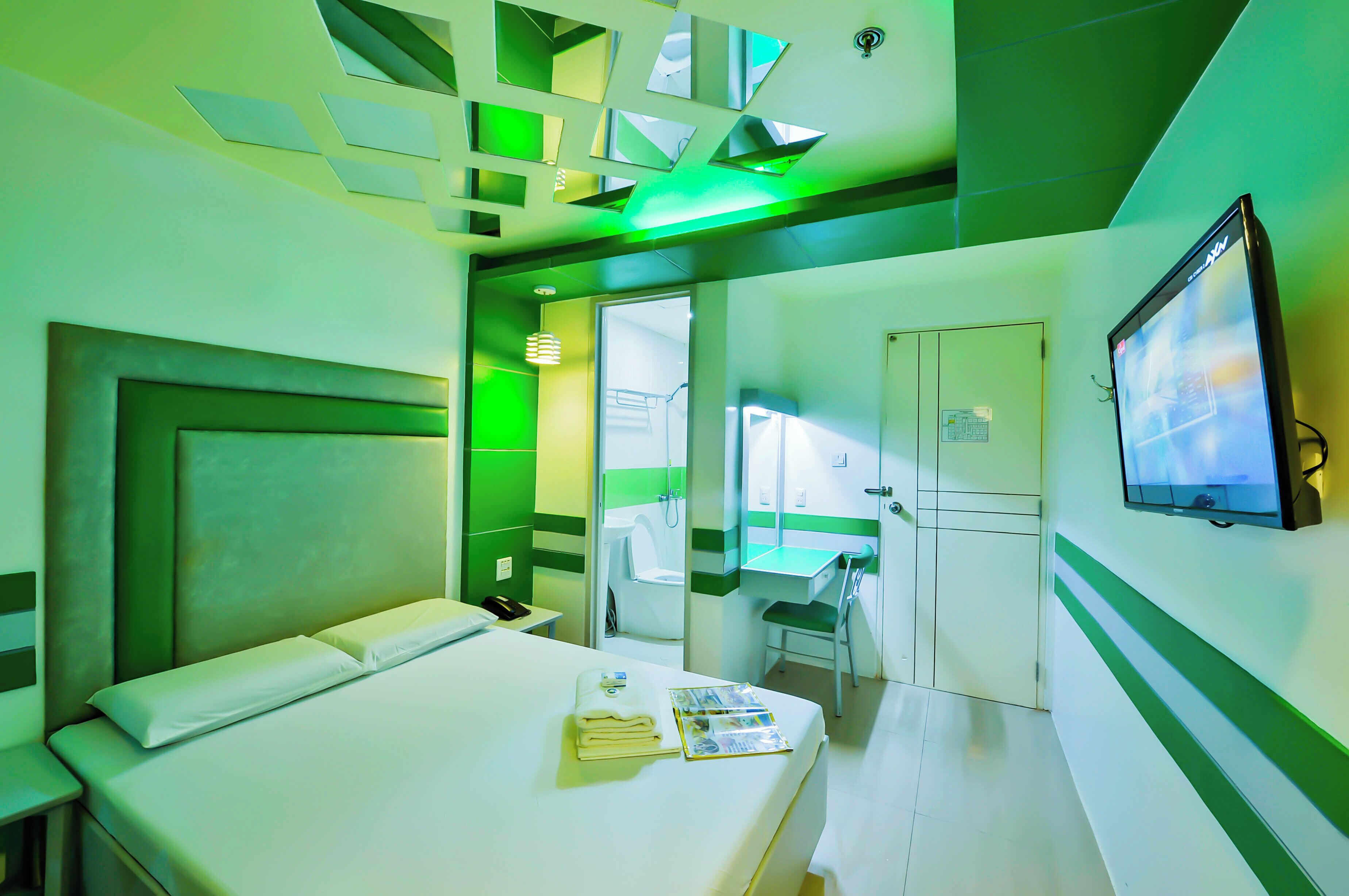 A hotel room with a bed, a bedside table, a mirror, a TV, and a bathroom visible through an open door.