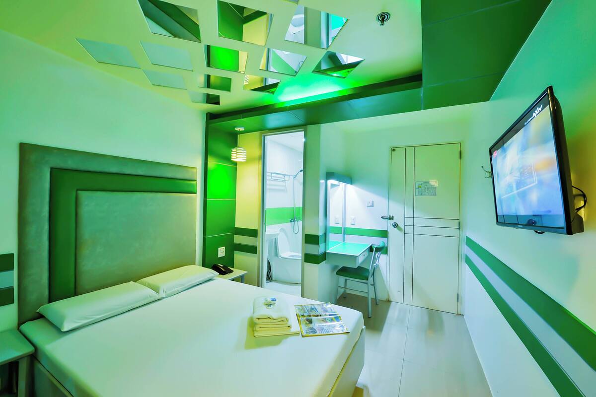 A hotel room with a bed, a bedside table, a mirror, a TV, and a bathroom visible through an open door.