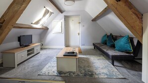 A loft bedroom with a sloped ceiling, a wooden beam structure, a television on a wooden console, a sofa with teal cushions, and a small table. - The Coach House (Corsham)