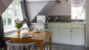 A dining area with a wooden table set for two, white chairs, a vase of flowers, and a kitchen in the background with white cabinets and a microwave. - The Coach House (Corsham)