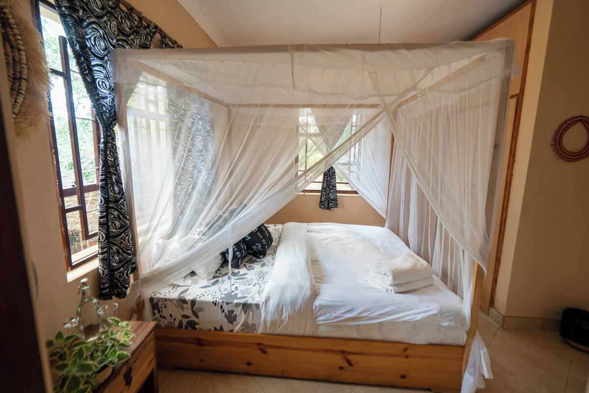 Ein Schlafzimmer mit einem hölzernen Bett, weißer Bettwäsche, einem Fenster mit gemusterten Vorhängen und einer Zimmerpflanze auf dem Nachttisch.