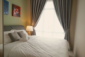 A neatly made bed with white linens and two pillows, a bedside lamp, and a view of the outdoors through curtains.