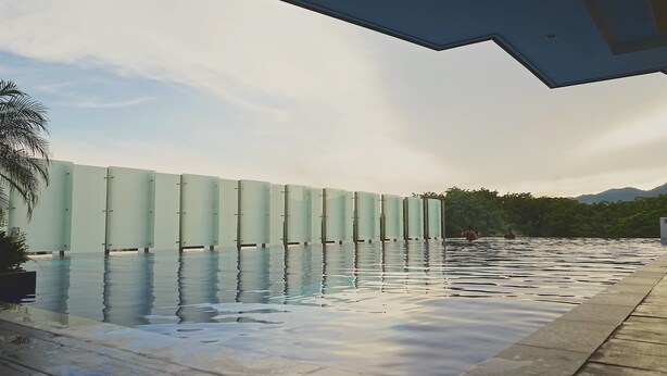 Piscina com parede de vidro, rodeada por um deck pavimentado e palmeiras.