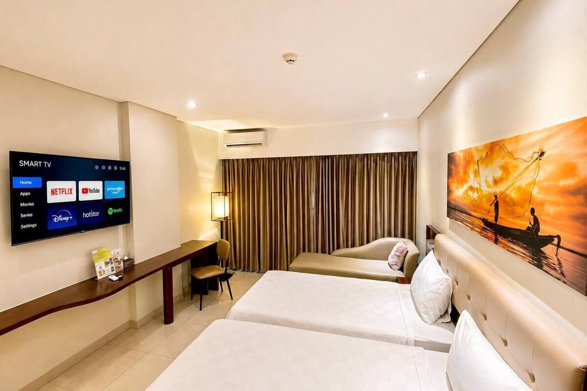 A hotel room with a large flat-screen TV displaying a smart TV interface, two beds, a desk with a chair, and a painting on the wall.