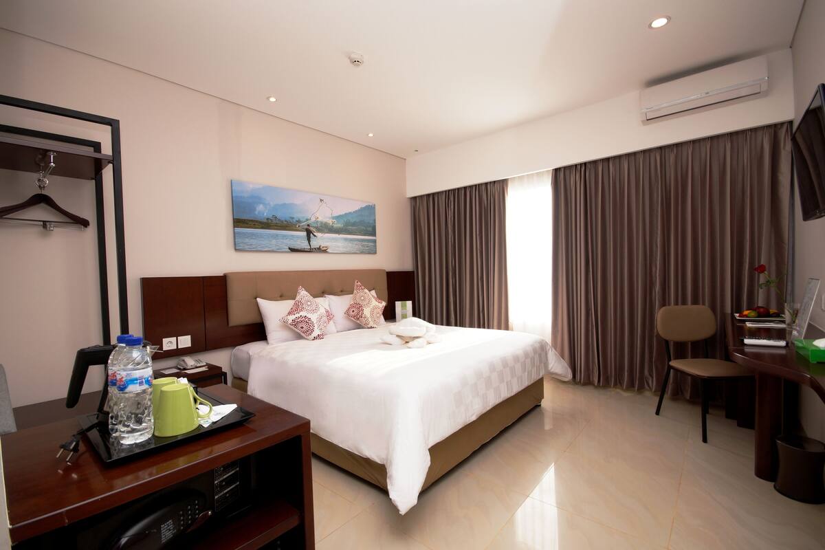 A hotel room with a large bed, a desk with a tray of toiletries, a chair, a desk lamp, and a painting on the wall.