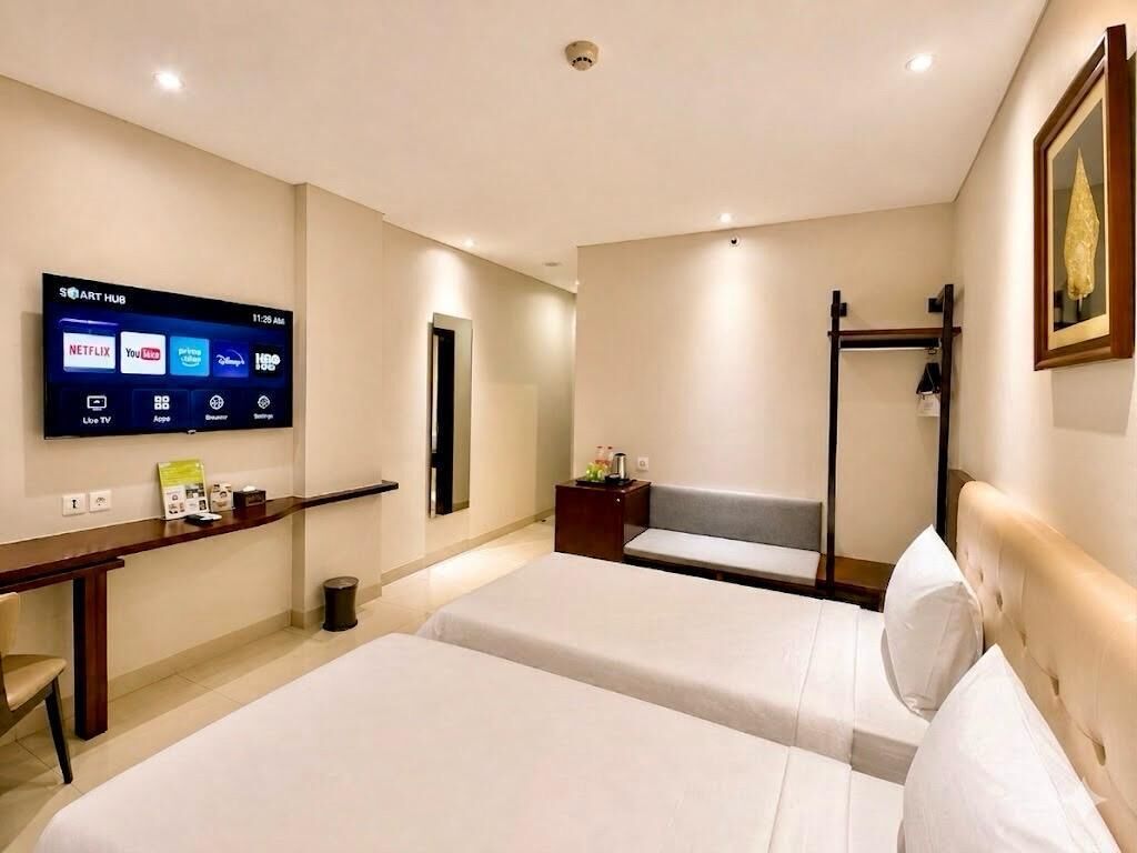 A hotel room with two beds, a TV displaying streaming services, a desk with a chair, and a bench.