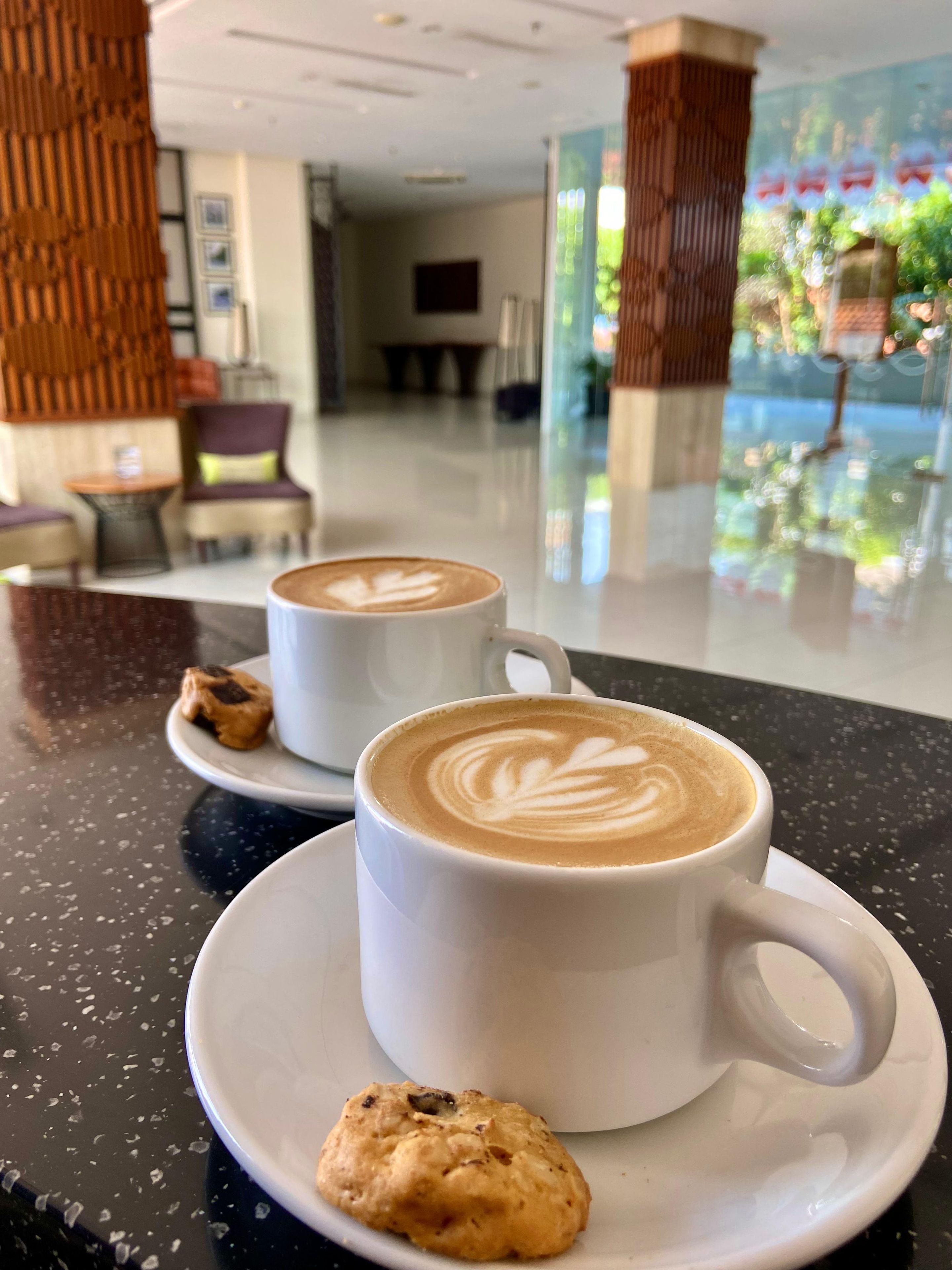 Dos tazas blancas de café con arte de latte sobre una encimera negra con motas, acompañadas de una galleta.