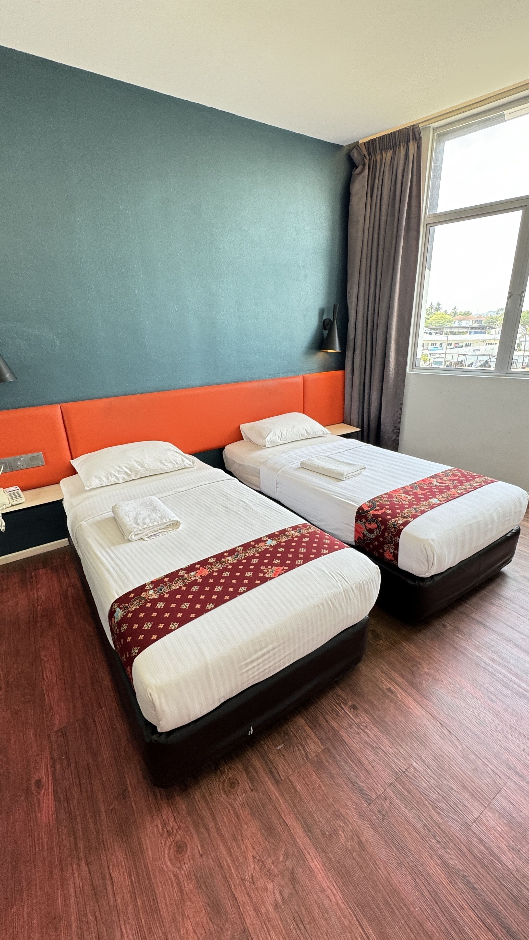 Habitación de hotel con dos camas, suelo de madera, pared azul y una ventana con cortinas.