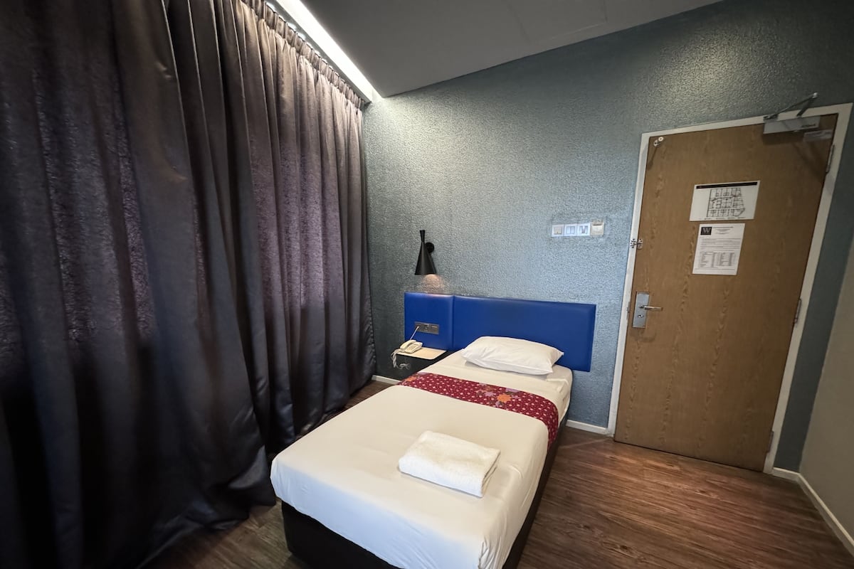 A hotel room with a bed, a nightstand, a lamp, a door, and a window with curtains.