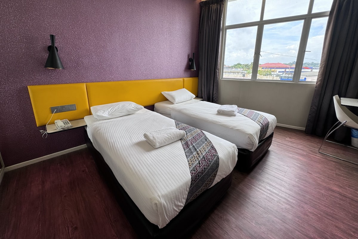 Habitación de hotel con dos camas, paredes moradas, cabecera amarilla, una ventana con vistas y un escritorio con silla.