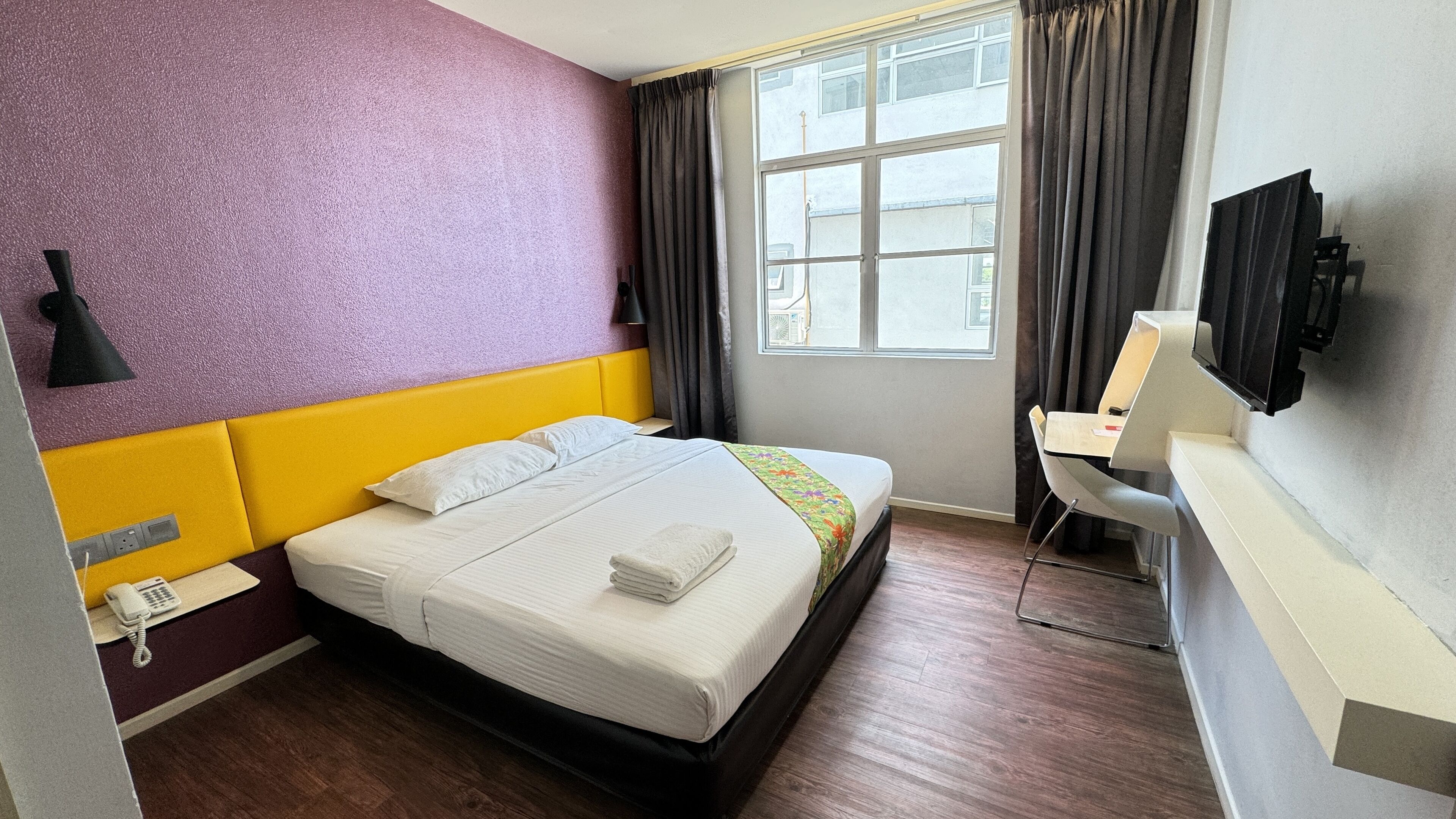 Habitación de hotel con cama, escritorio, silla, televisor, teléfono y ventana con cortinas.