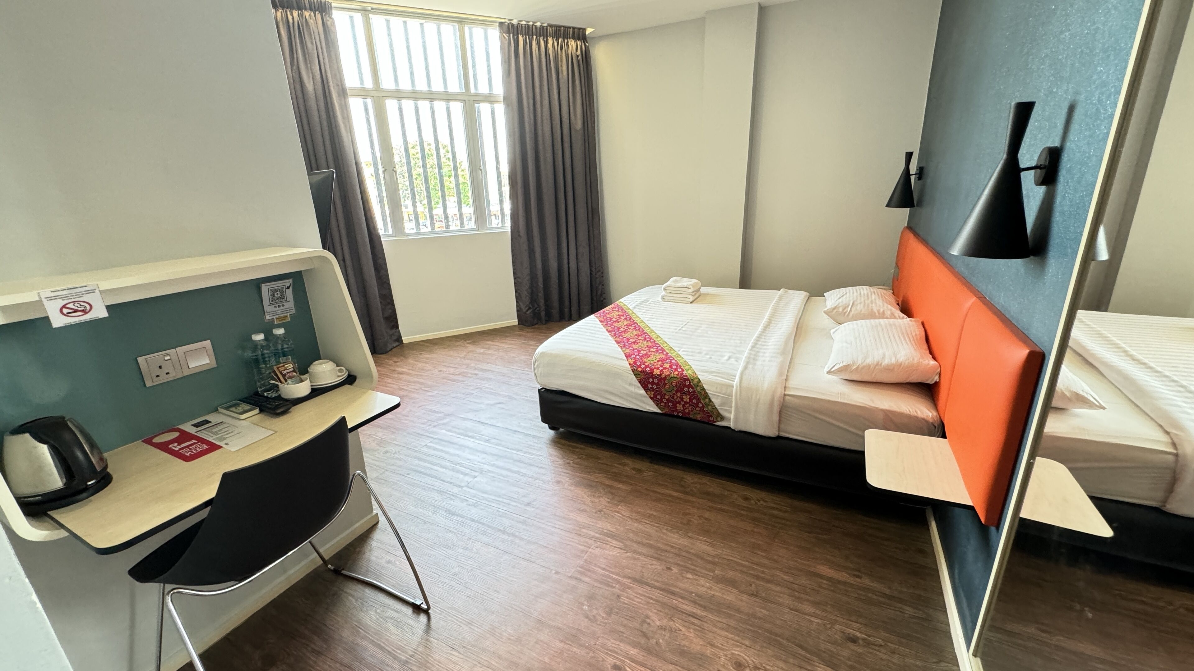 Una habitació d'hotel amb un llit, una taula de treball amb una cadira, una làmpada de taula i una finestra amb cortines.