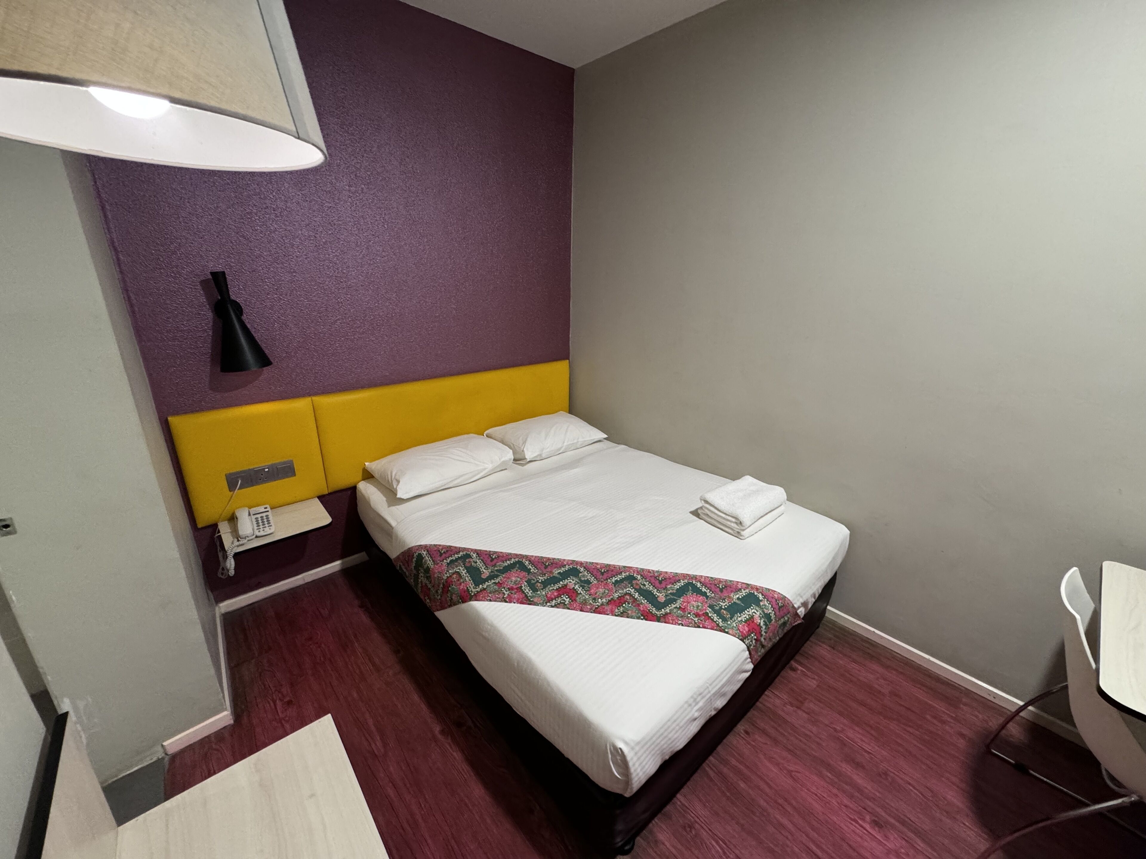 A hotel room with a bed, a bedside table with a lamp, a phone, and a chair.