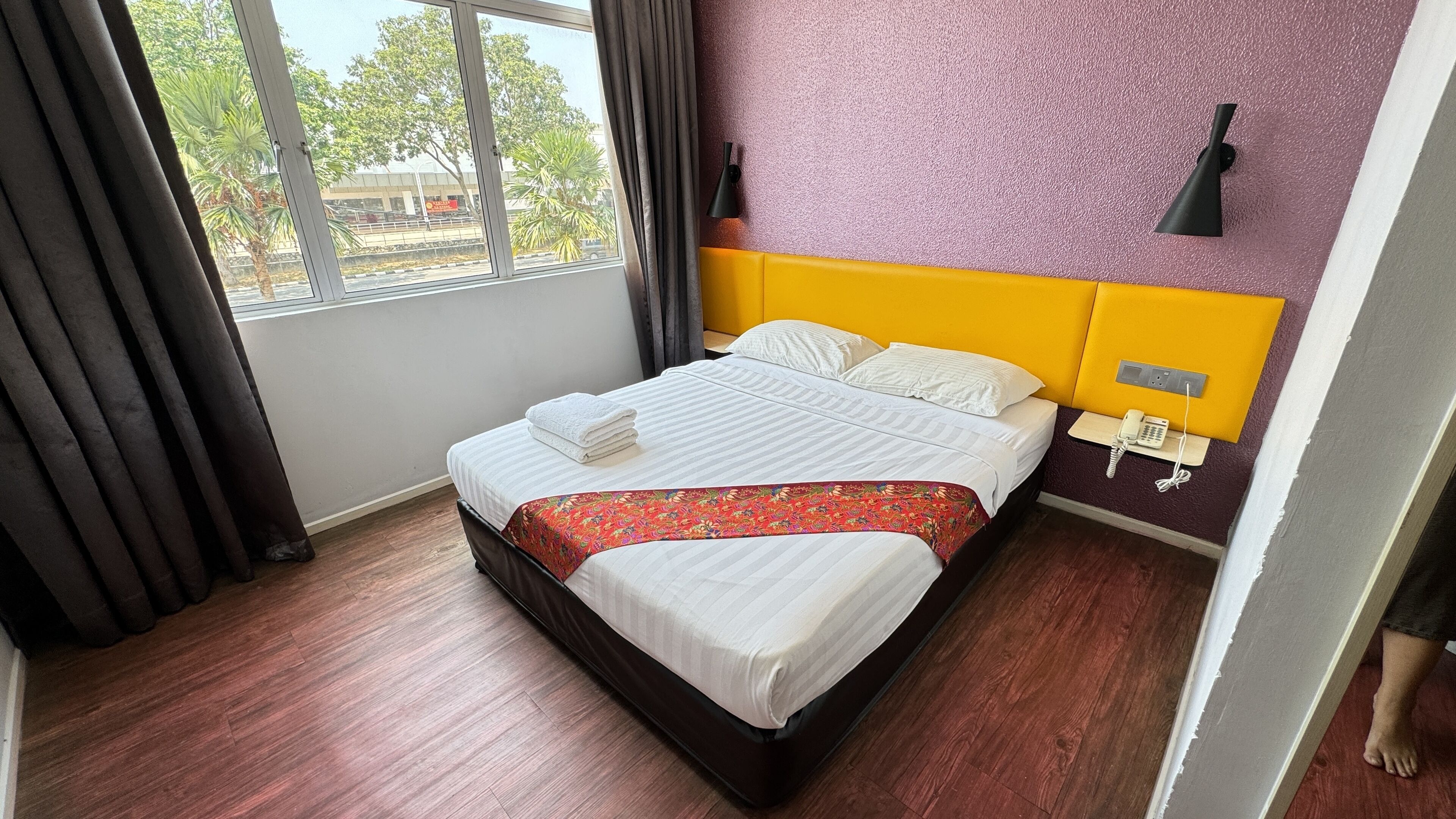 A hotel room with a bed, a bedside telephone, and a window with curtains.