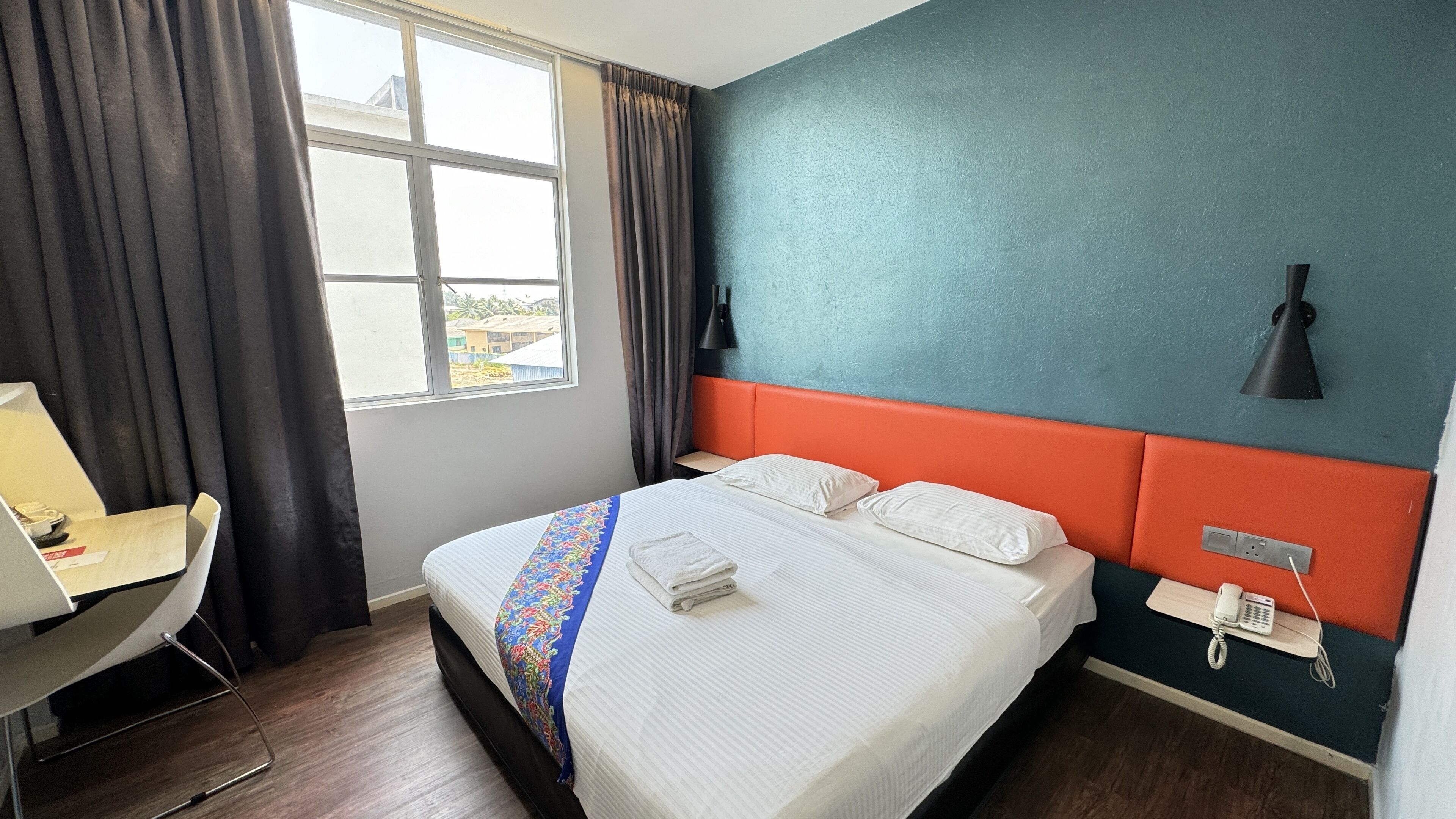 A hotel room with a bed, a desk, a chair, a window with curtains, and a wall-mounted phone.