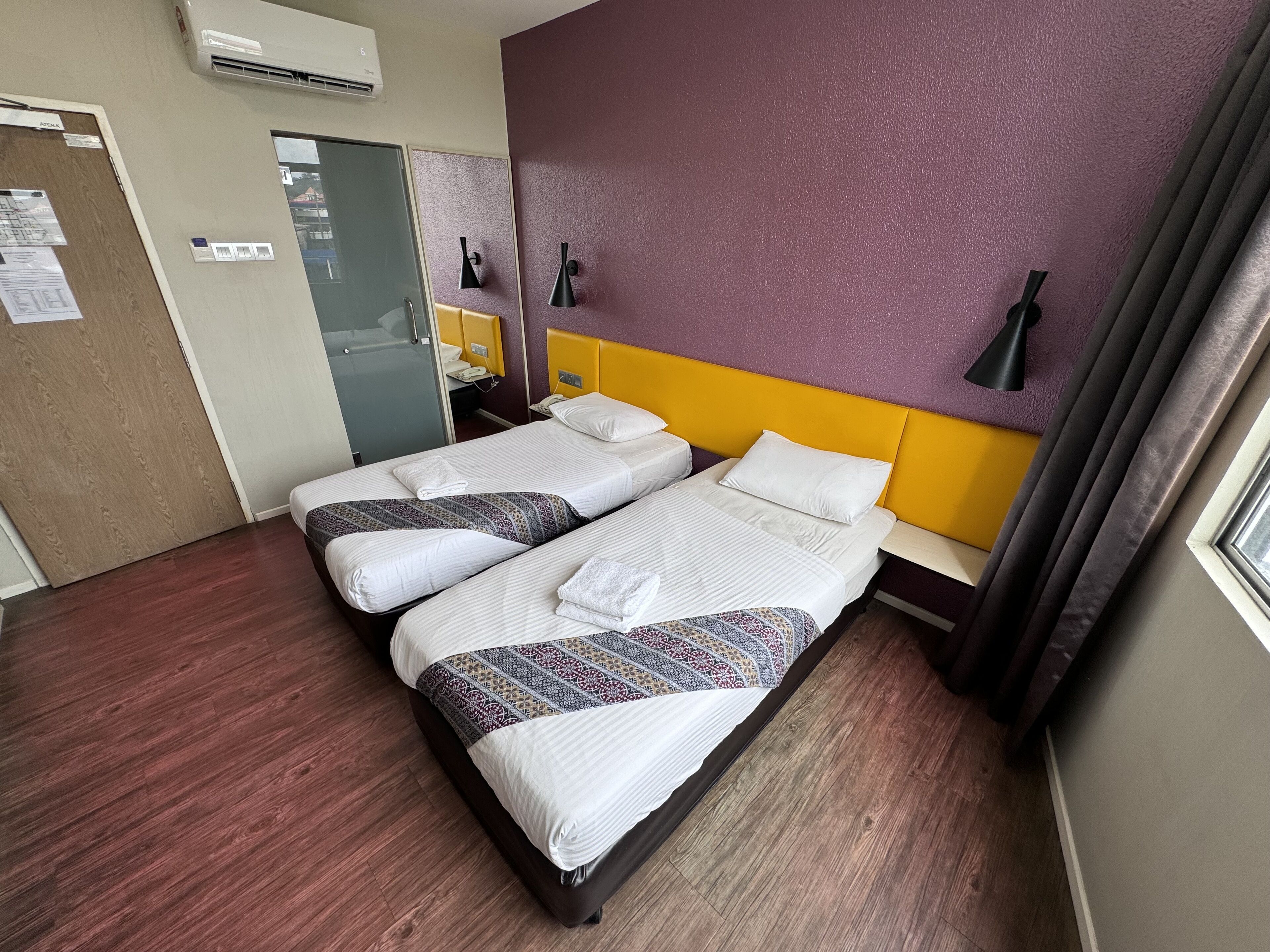 Une chambre d’hôtel avec deux lits, un plancher en bois, un mur d’accent violet et une fenêtre avec des rideaux.