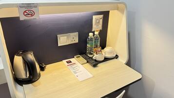 A hotel room with a desk, a coffee maker, a microwave, a water bottle, a coffee cup, and a QR code for hotel services.