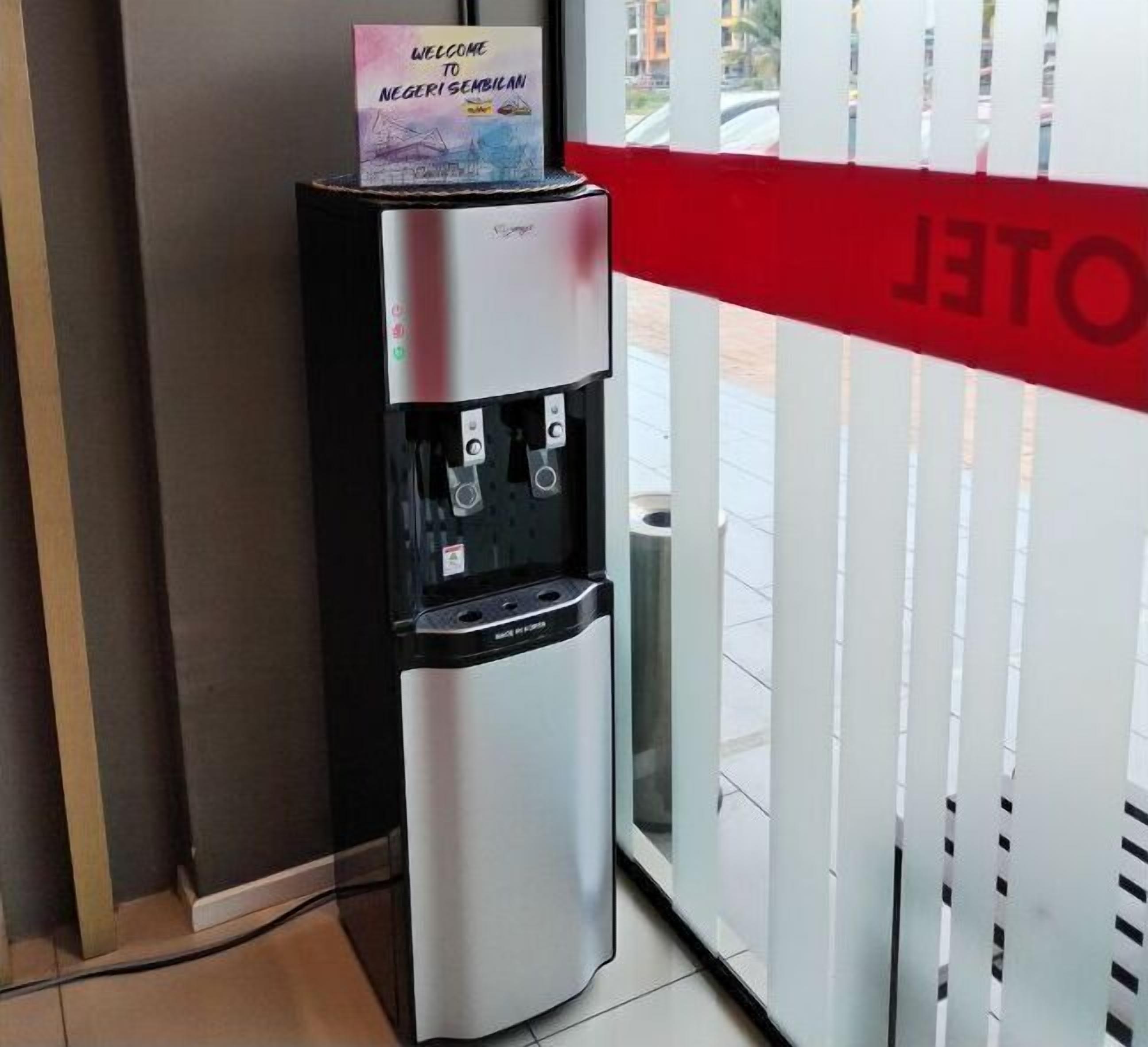 Un dispensador de agua con un cartel de bienvenida, ubicado cerca de un vidrio con un letrero rojo.