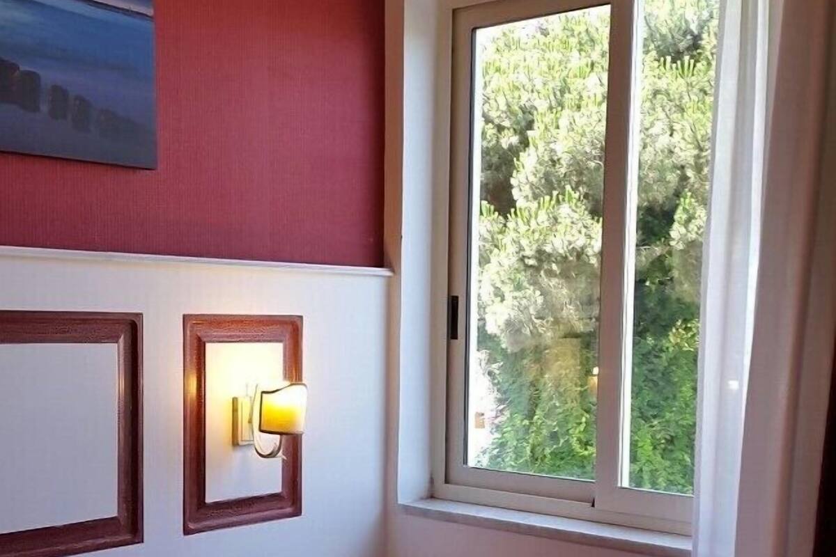 A hotel room with a bed, a small bedside table, a wall-mounted light, and a window with a view of greenery.